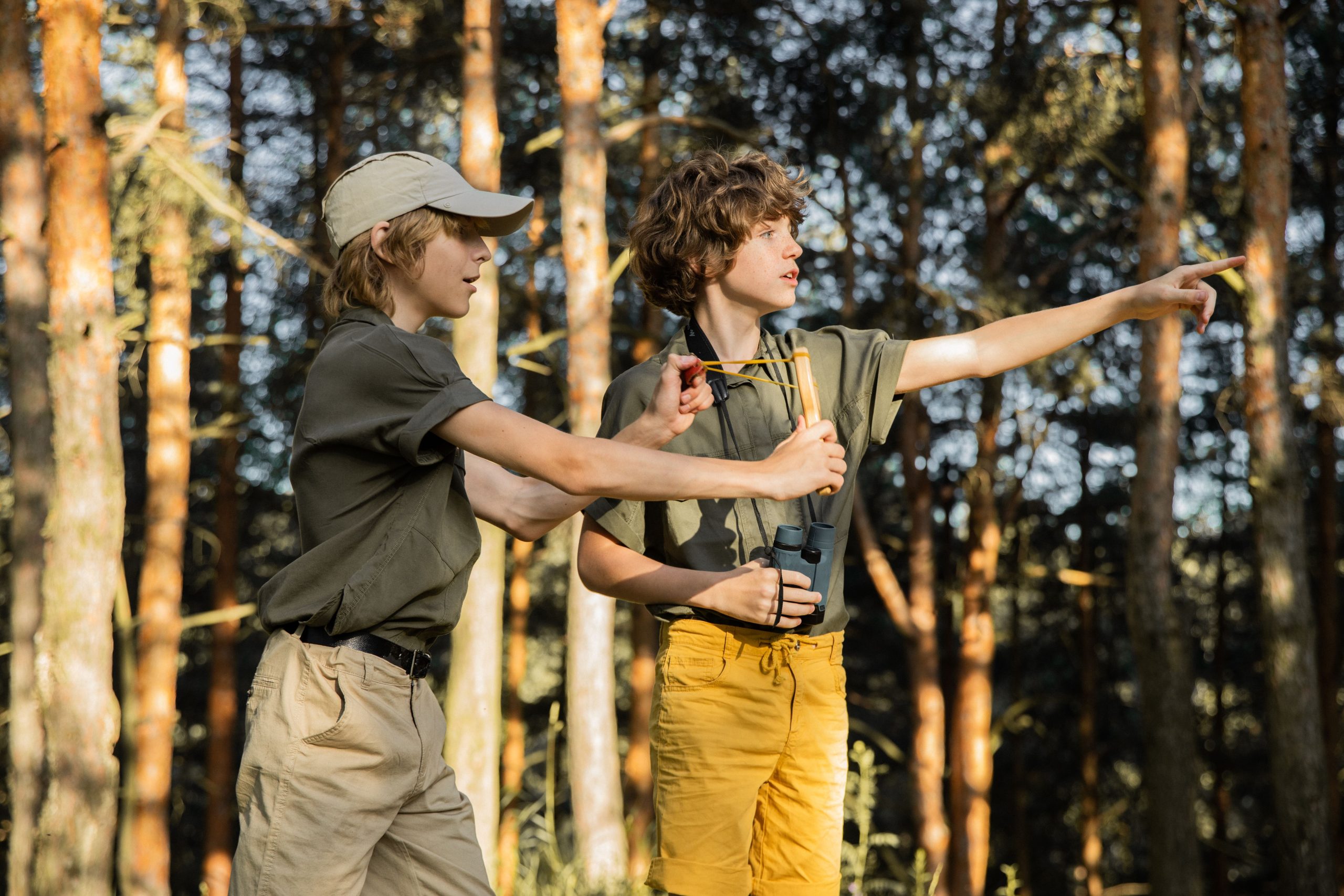 Two teenage boys in forest using slingshot and pointing, embodying adventure and exploration.